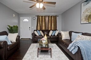 A living room with two brown leather sofas, a glass coffee table, a white rug, a ceiling fan, and a door with curtains. - Cozy 3-bedroom Duplex1 with WiFi, AC in peaceful Springfield (SPRINGFIELD)