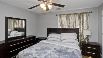 A bedroom with a bed, a ceiling fan, a nightstand, a dresser, a mirror, and a window with curtains.