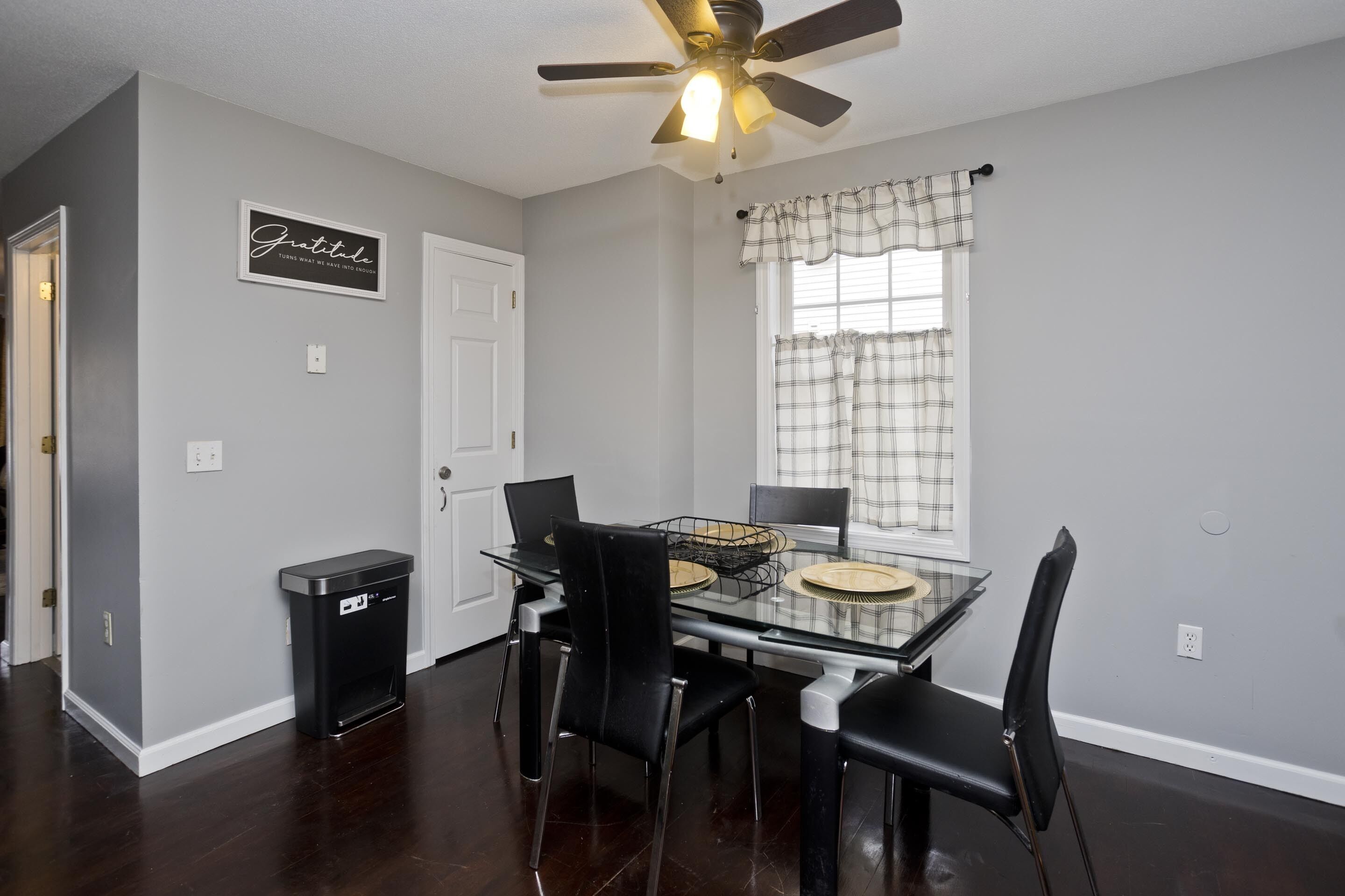 A dining area with a glass table, black chairs, a ceiling fan, and a window with plaid curtains.