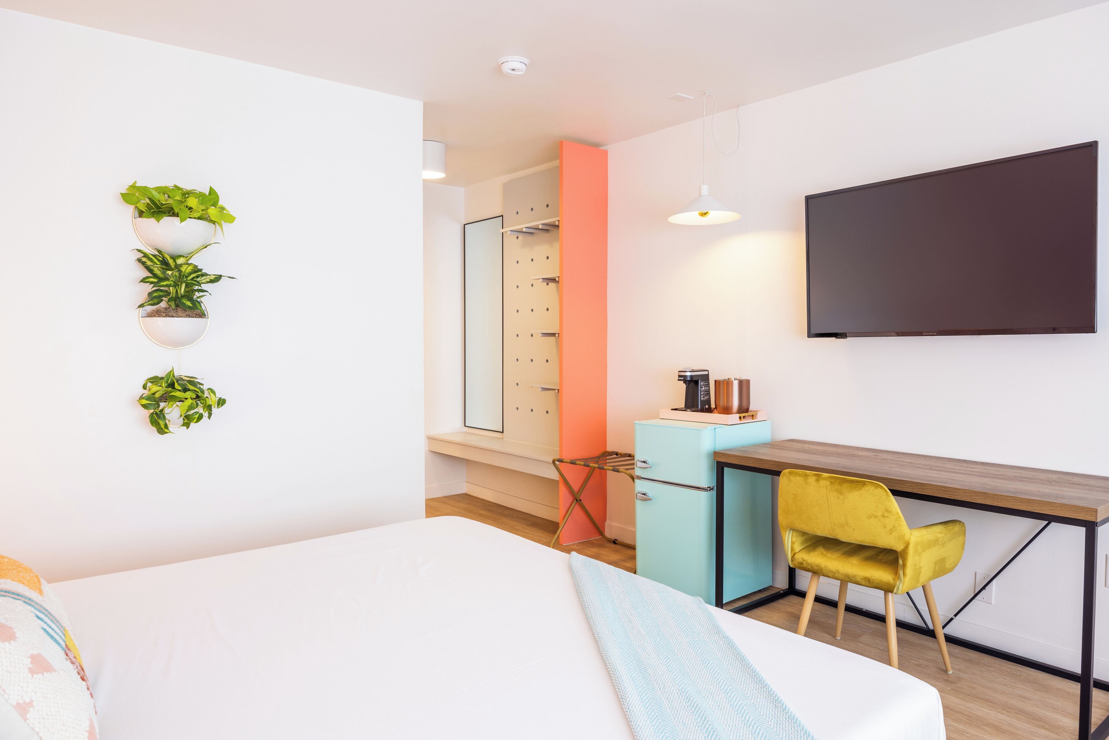 Moderni hotellihuone, jossa on sänky, työpöytä, tuoli, televisio ja seinälle kiinnitetyt kukkaruukut.