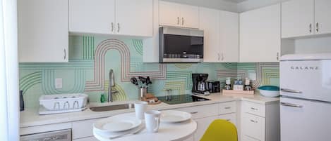 A modern kitchen with white cabinets, a microwave, a coffee maker, and a refrigerator. There is a dining table with chairs and a sink with a faucet.