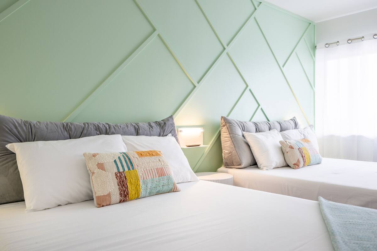 A hotel room with two beds, each with white linens and multiple pillows. The room features a green accent wall with a geometric pattern and a white ceiling.