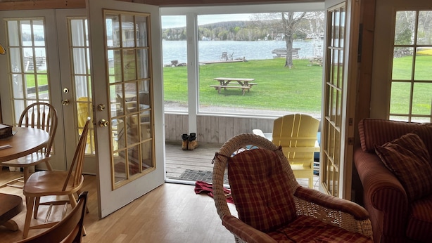 A room with a wooden interior, a dining area with chairs and a table, and a view of a lake with a picnic table outside. - Lake Willoughby Direct Waterfront (Westmore)