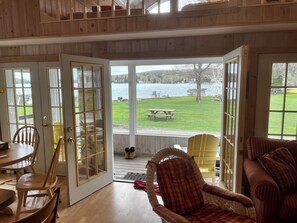 A room with a wooden interior, a dining area with chairs and a table, and a view of a lake with a picnic table outside. - Lake Willoughby Direct Waterfront (Westmore)
