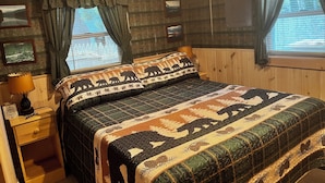 A cozy bedroom with a bed, wooden walls, and a window with curtains. - Lake Willoughby Direct Waterfront (Westmore)