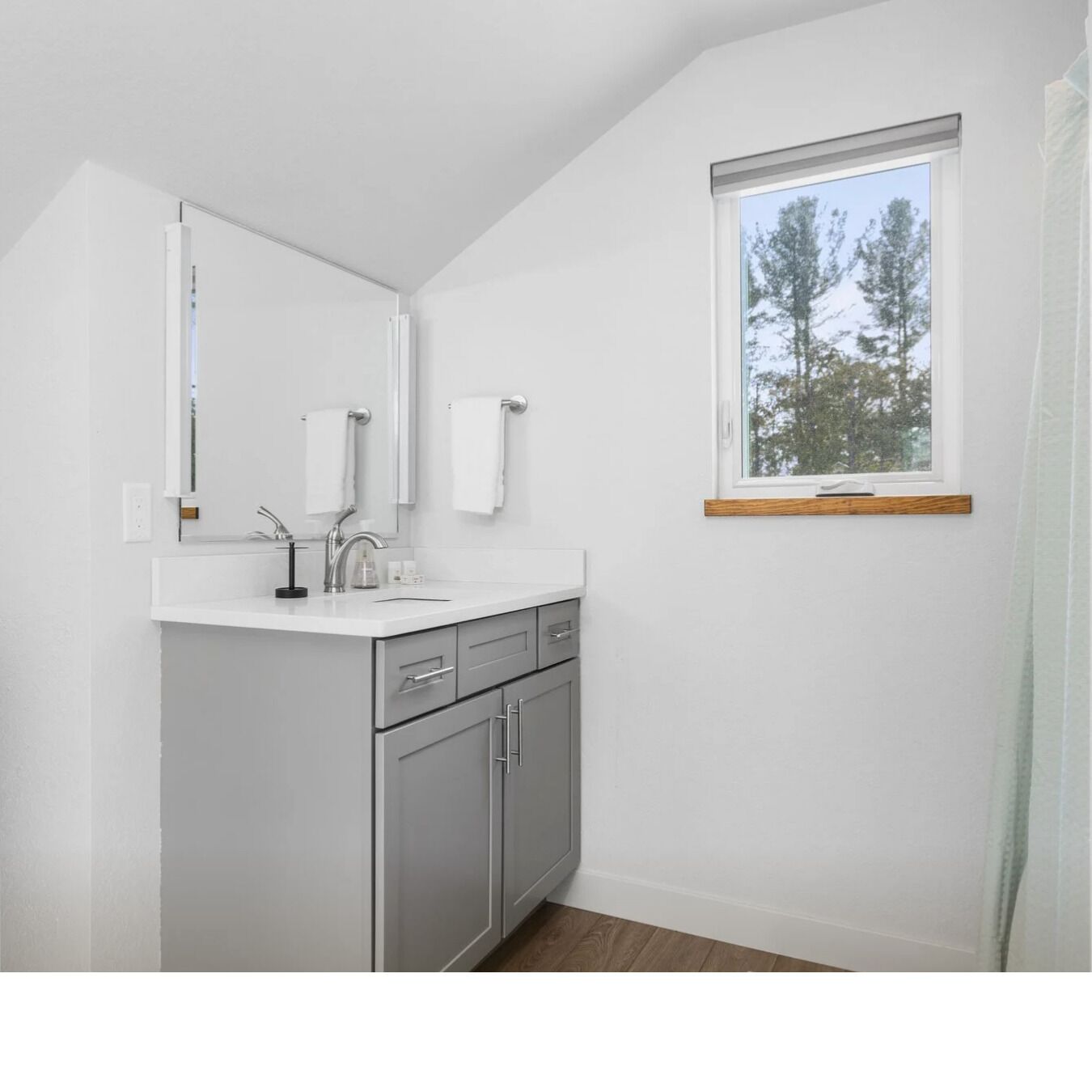 Un baño moderno con un tocador blanco, gabinetes grises, un espejo grande y una ventana con vista a árboles.