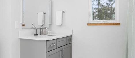 Un baño moderno con un tocador blanco, gabinetes grises, un espejo grande y una ventana con vista a árboles.