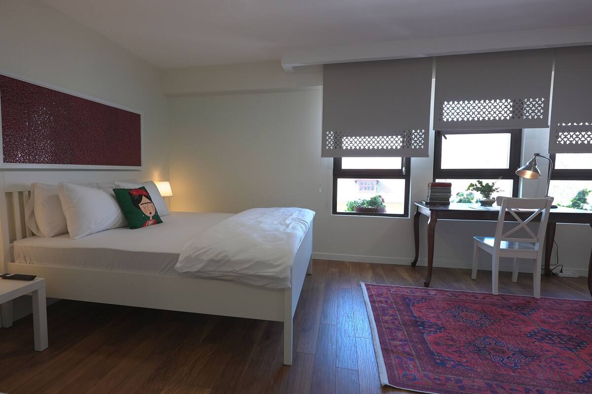 A bedroom with a bed, bedside table, lamp, desk, chair, and a view of the outside through a window with a lattice screen.