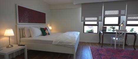 Quarto com cama, mesa de cabeceira, abajur, escrivaninha, cadeira e vista para o exterior através de uma janela com grade.