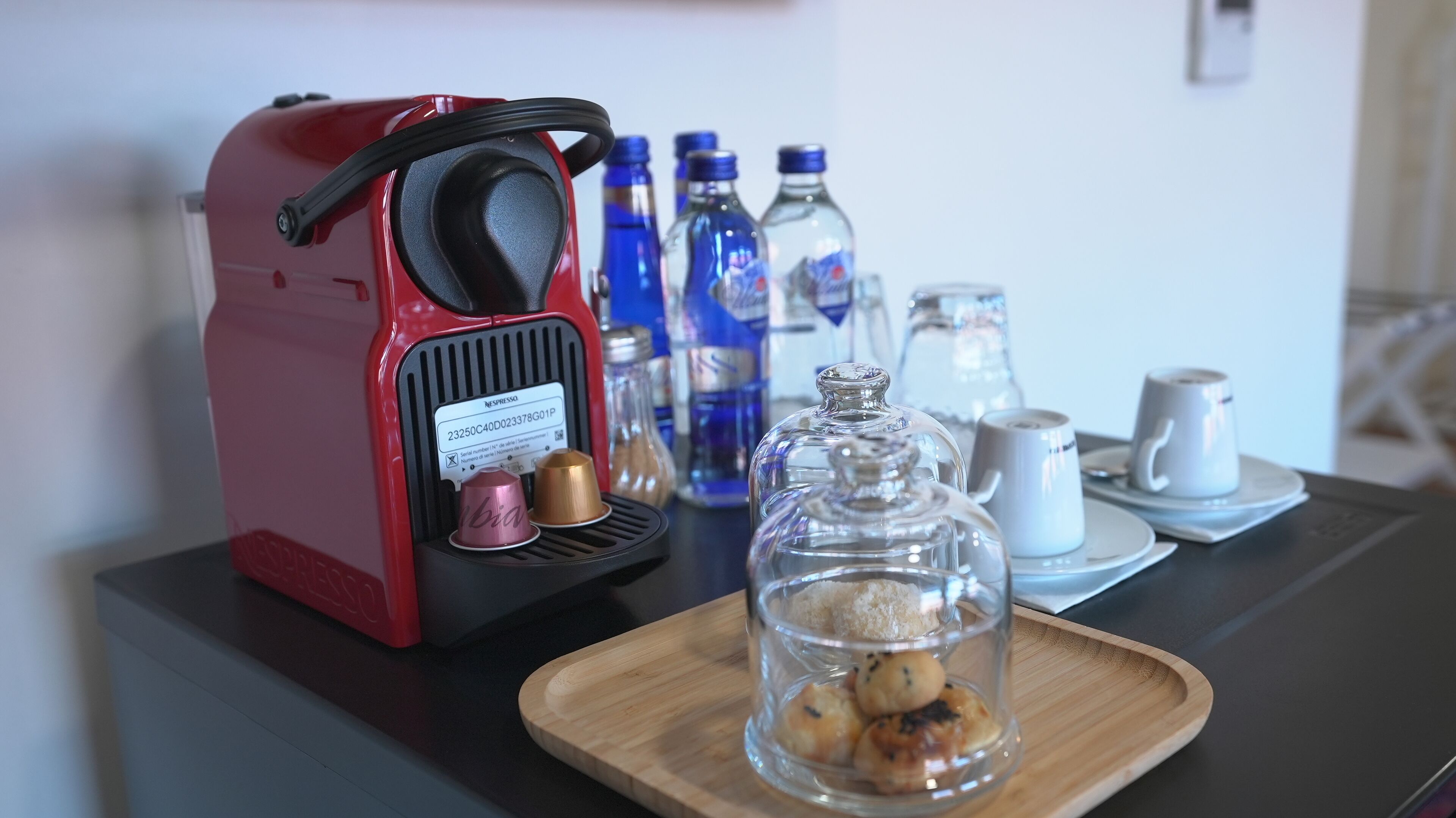 A red Nespresso coffee machine on a table with a glass jar of cookies, a white mug, and blue water bottles.