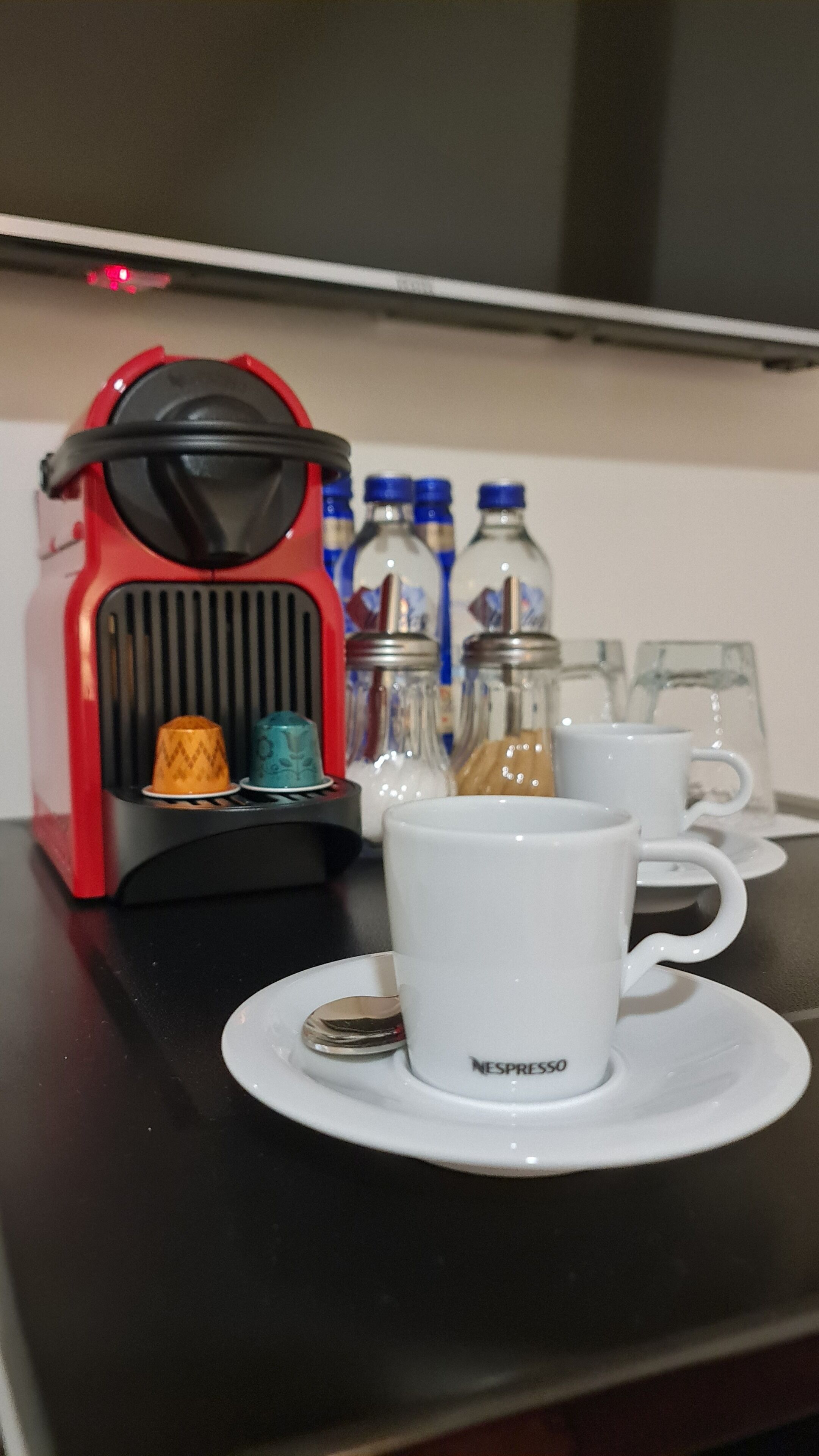 Eine Kaffeemaschine mit rotem und schwarzem Design, eine weiße Tasse mit 'NESPRESSO', sowie mehrere Glasflaschen und Tassen auf einem Tisch.