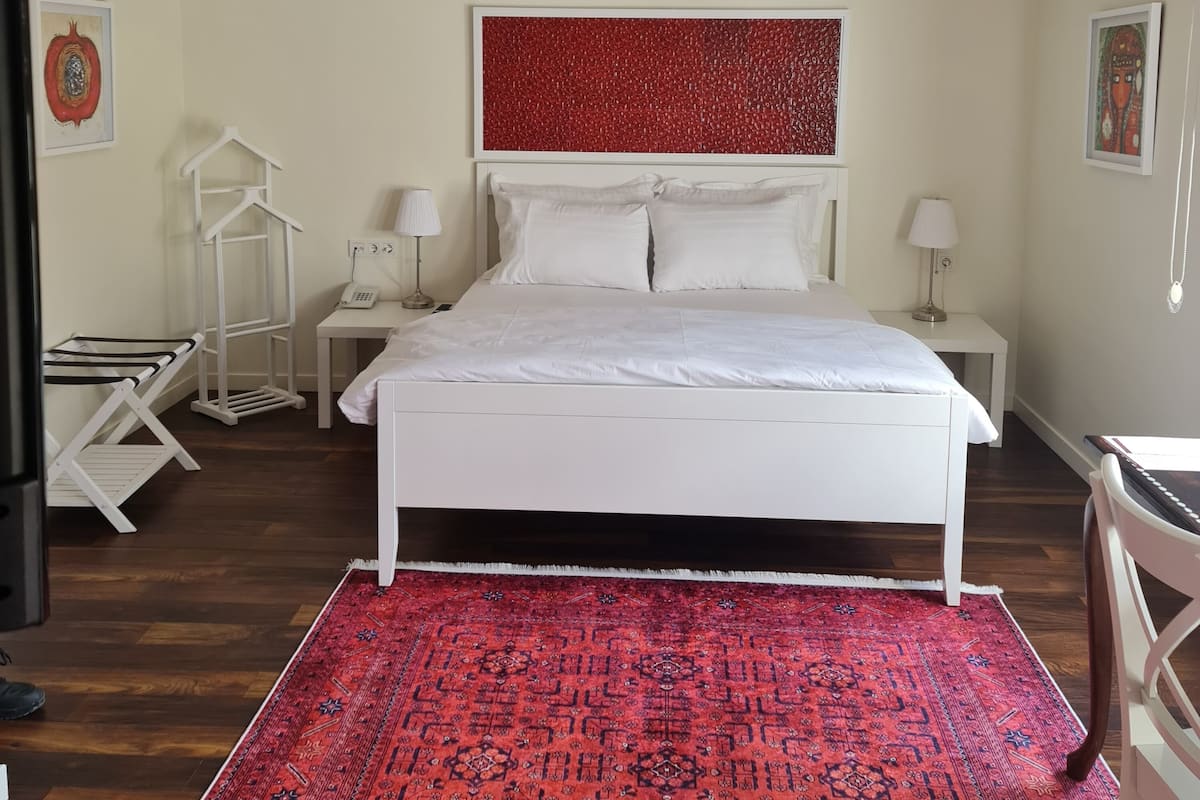 A bedroom with a white bed, a red patterned rug, a white nightstand with a lamp, and a white wardrobe.