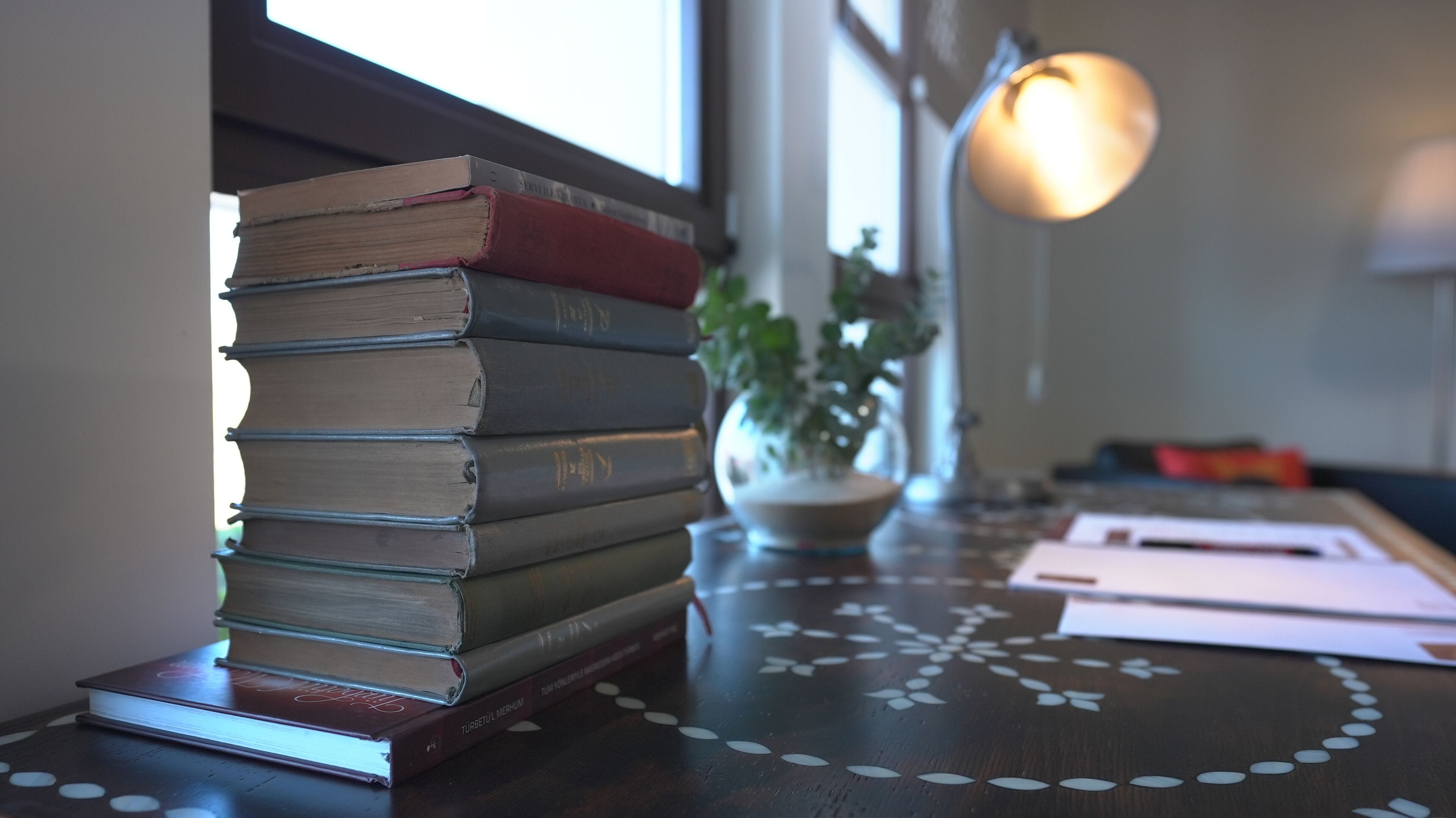 A stack of books on a wooden table with a decorative pattern, a potted plant, and a lamp in a room with a window.