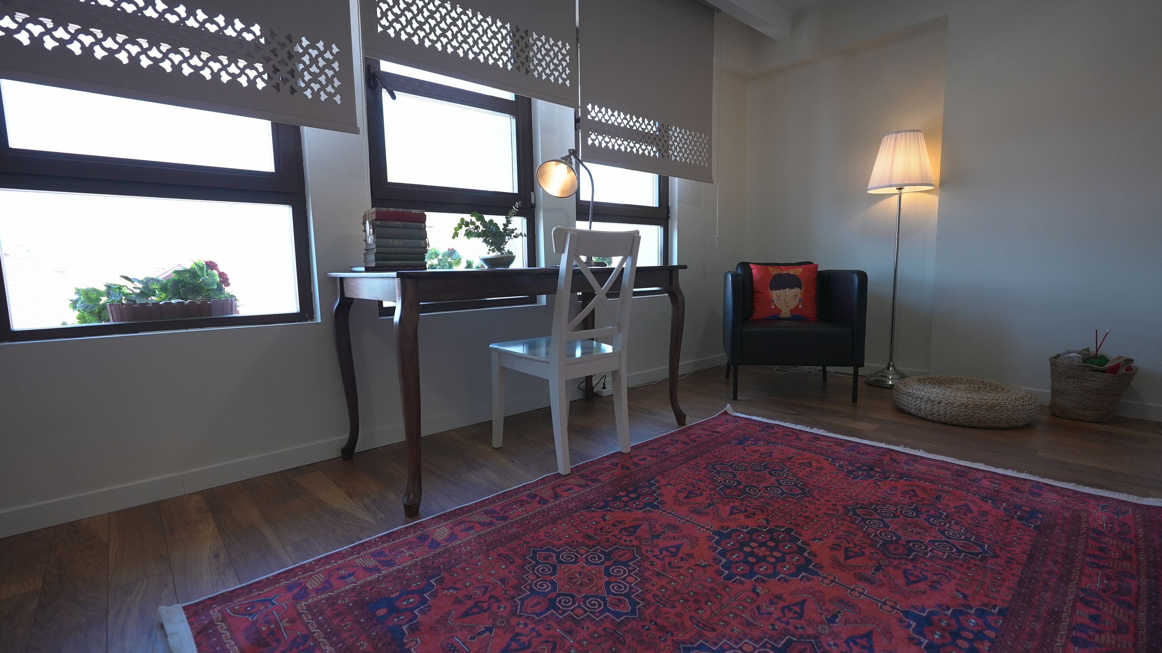 Una habitación con un ventanal grande, un escritorio con libros, una silla blanca, una silla negra con cojín, una lámpara de pie y un tapete rojo estampado.