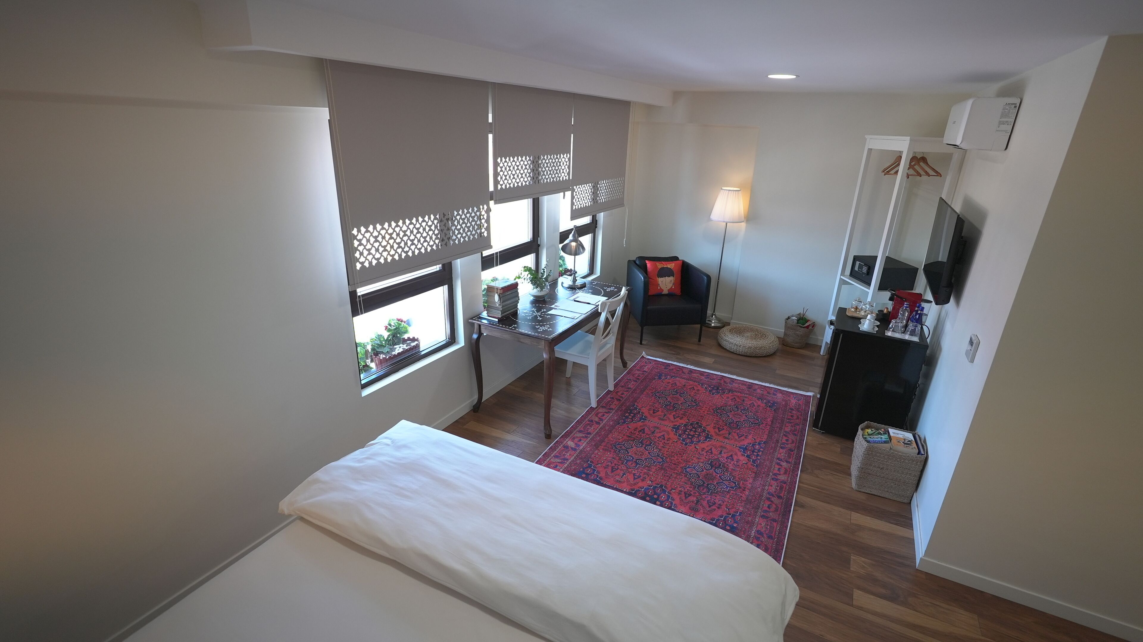 A bedroom with a bed, a desk with a chair, a small table with a lamp, a sofa, a TV, and a window with a view of plants outside.
