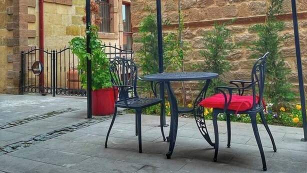 Une terrasse extérieure avec un bâtiment en pierre, des chaises en métal, une table et des auvents rouges.