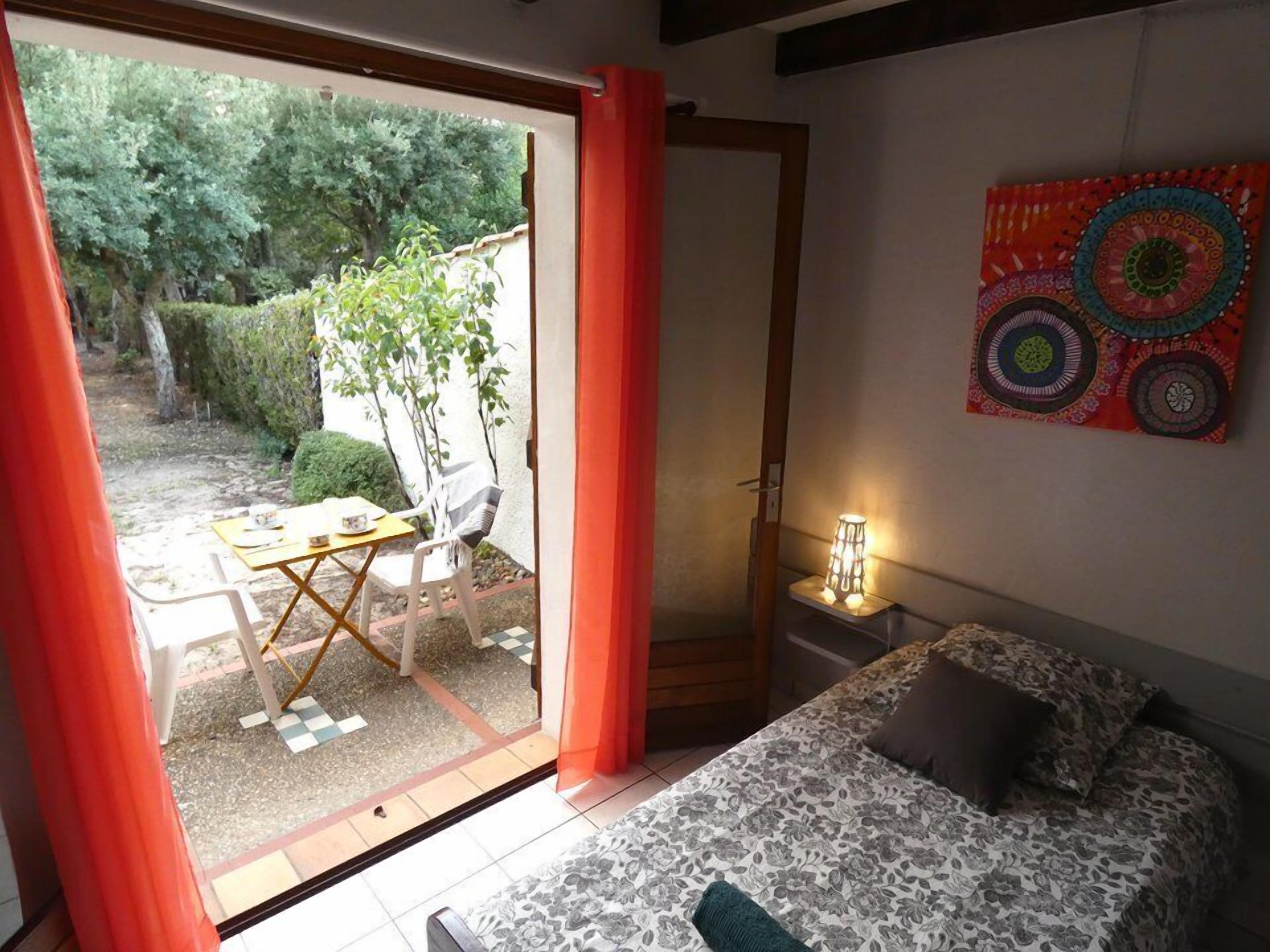 Une chambre à coucher comprenant un lit, une table de chevet, une table avec des chaises, et donnant sur le jardin grâce à une porte ouverte.