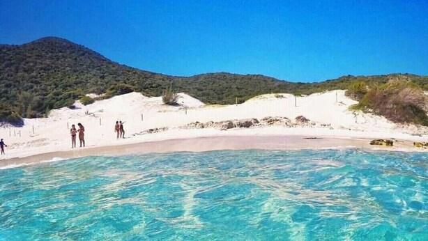 A clear blue pool with a sandy beach and a hilly landscape in the background. - Prédio Atalaia  (Arraial do Cabo)