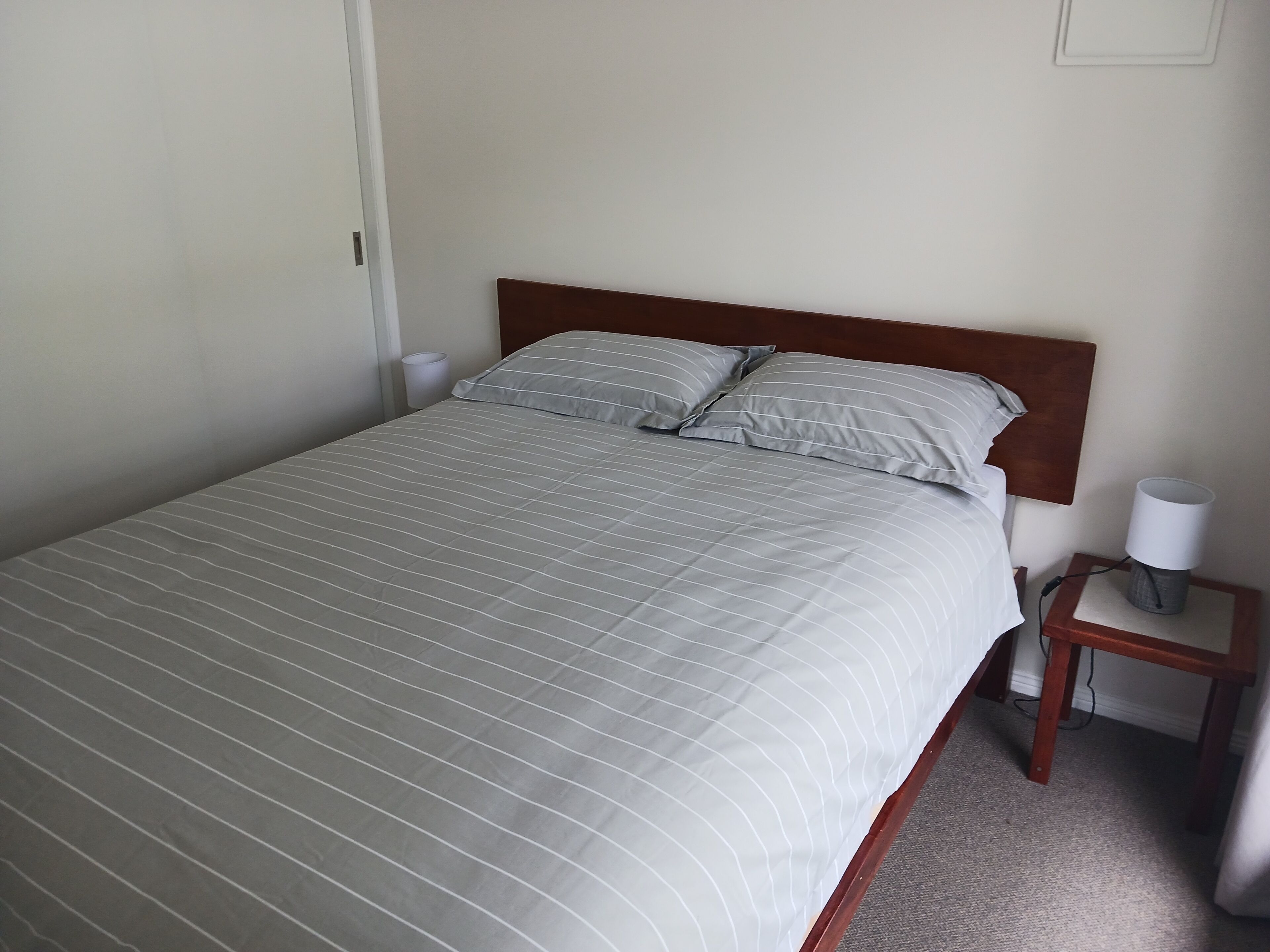 A neatly made bed with a striped bedspread and two pillows, a wooden headboard, a bedside table with a lamp, and a wall-mounted light fixture.