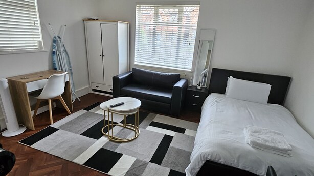 A modern bedroom with a wooden herringbone floor, a black and white striped rug, a black sofa, a white bed with a white pillow, a small round table, a white wardrobe, and a window with blinds.