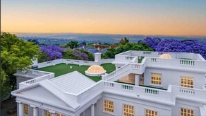Un bâtiment blanc doté de nombreux balcons et bordé d’une pelouse verdoyante, entouré d’arbres et de fleurs violettes.