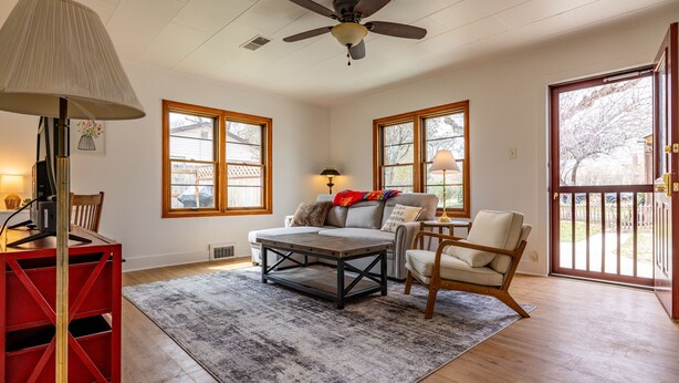 A spacious living room with a ceiling fan, two windows with wooden frames, a red metal TV stand, a grey sofa, a wooden coffee table, and a patterned rug.