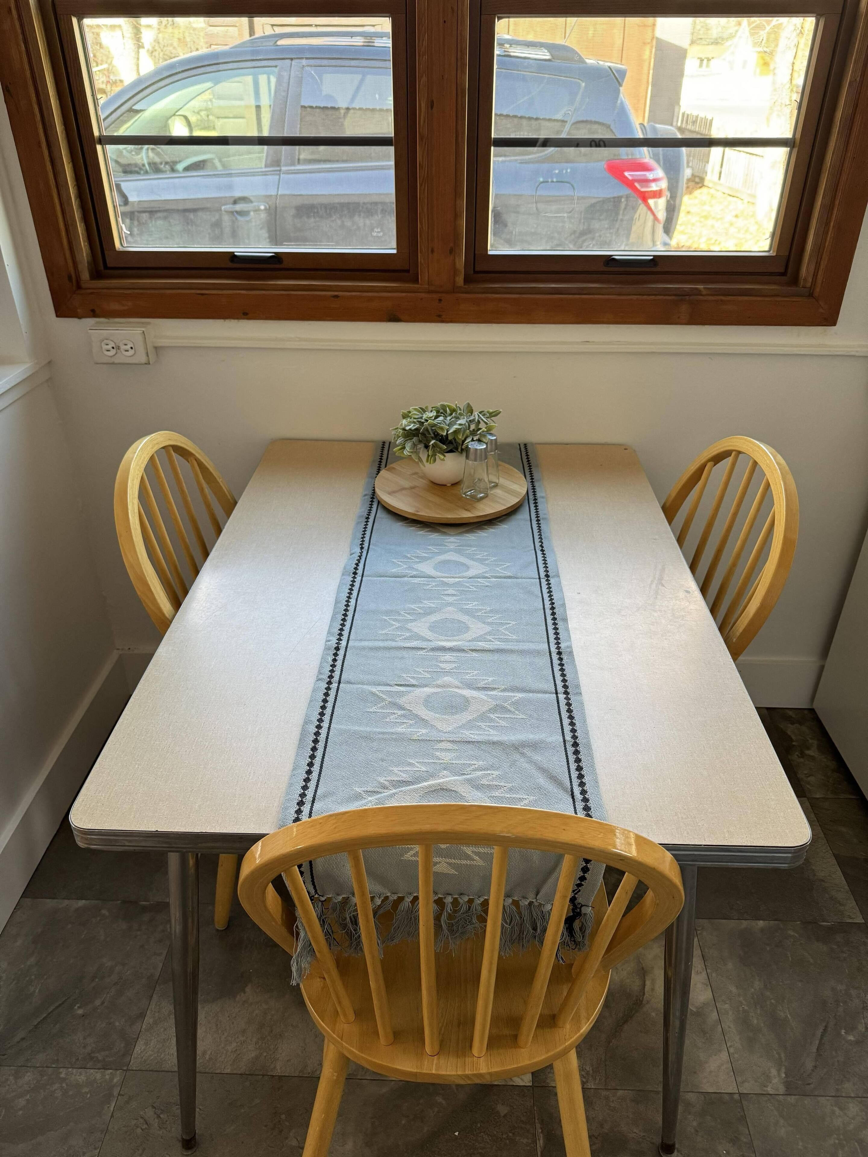 A dining table with a blue patterned runner, two wooden chairs, and a small potted plant on a wooden tray.
