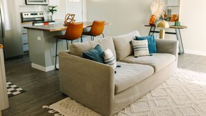 Ein modernes Wohnzimmer mit einem beigefarbenen Sofa, einem Essbereich mit orangefarbenen Stühlen und einer Küche mit weißen Schränken und einer Mikrowelle.