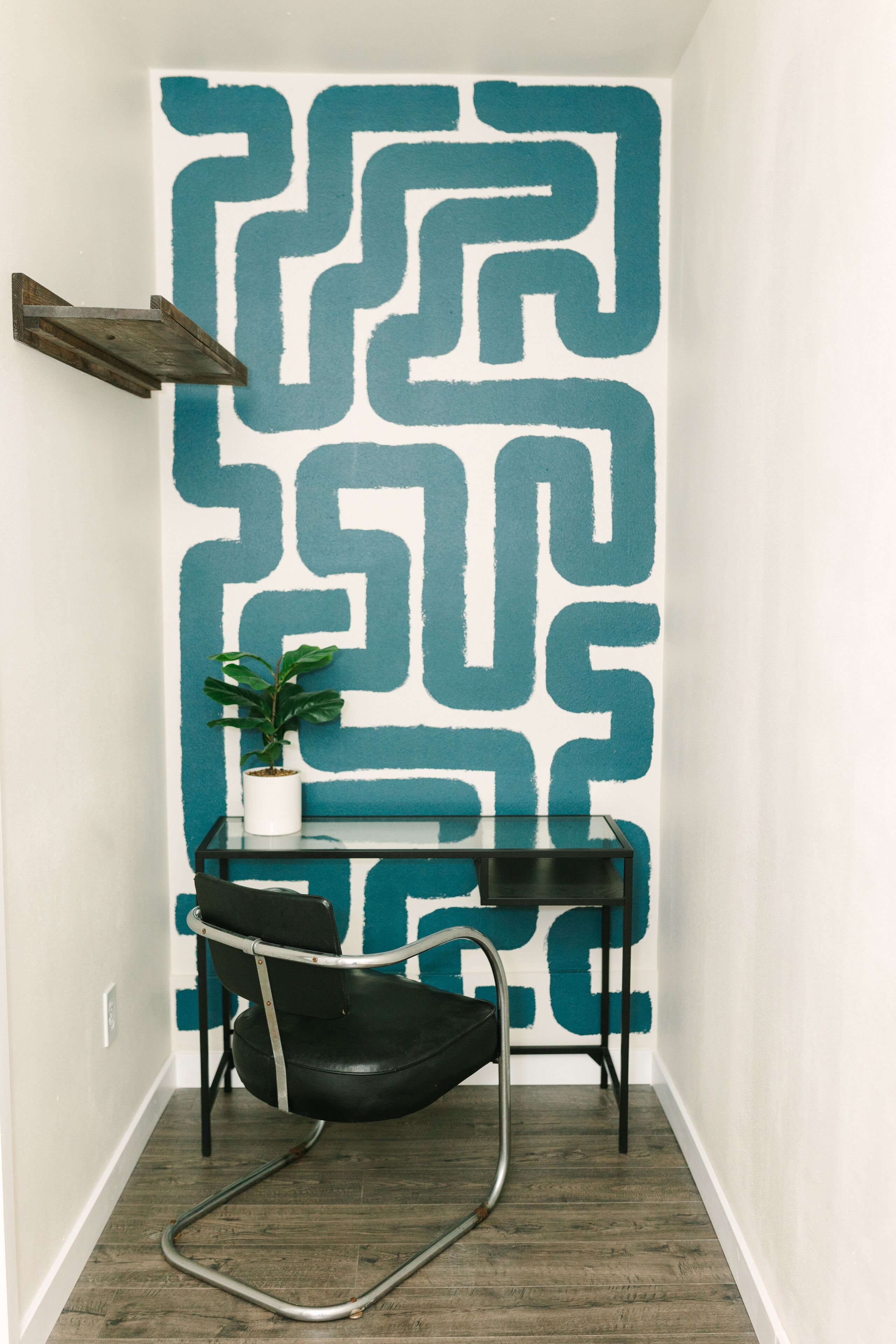 A narrow hallway with a geometric wall mural, a black chair, a small table with a potted plant, and a wooden shelf.