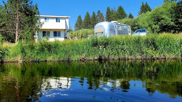 A house with a white exterior, a trailer, and a car parked near a body of water with reflections. - Cozy cottage with river access and great outdoor adventures (La Pine)
