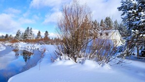 A snow-covered landscape with a house by a frozen pond, surrounded by trees and a clear sky. - Cozy cottage with river access and great outdoor adventures (La Pine)