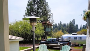 A wooden deck with outdoor furniture, a covered seating area, and a view of a forested area with a cabin and a trailer. - Cozy cottage with river access and great outdoor adventures (La Pine)