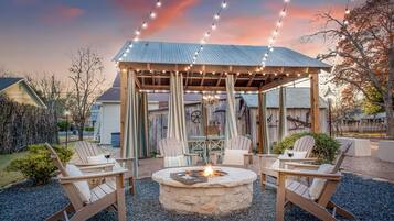A gazebo with a stone fire pit and seating, surrounded by a gravel area and string lights.