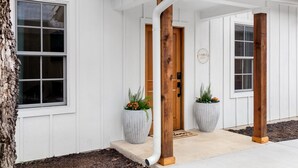 Una casa blanca con puerta de madera, dos macetas grandes y una placa identificadora visible.