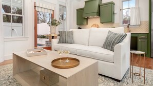 Una sala de estar moderna con un sofá blanco, una mesa de centro con bandeja y vista a la cocina con gabinetes verdes.
