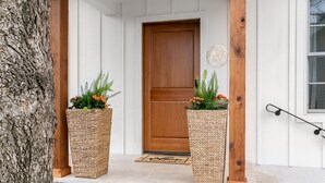 A wooden door with a doormat and two woven planters with flowers on a white porch. - Bijou: Collette | Luxury Cottage | Plunge Pool | Full Kitchen | Seasonal Fireplace (Fredericksburg)