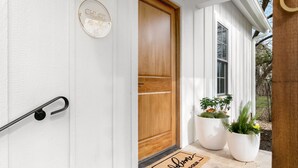 A white exterior with a wooden door, a nameplate, and two potted plants on the porch. - Bijou: Chloe | Luxury Cottage | Plunge Pool | Full Kitchen | Seasonal Fireplace (Fredericksburg)