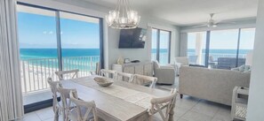 A spacious room with a large dining table, white furniture, and a balcony with a view of the ocean.