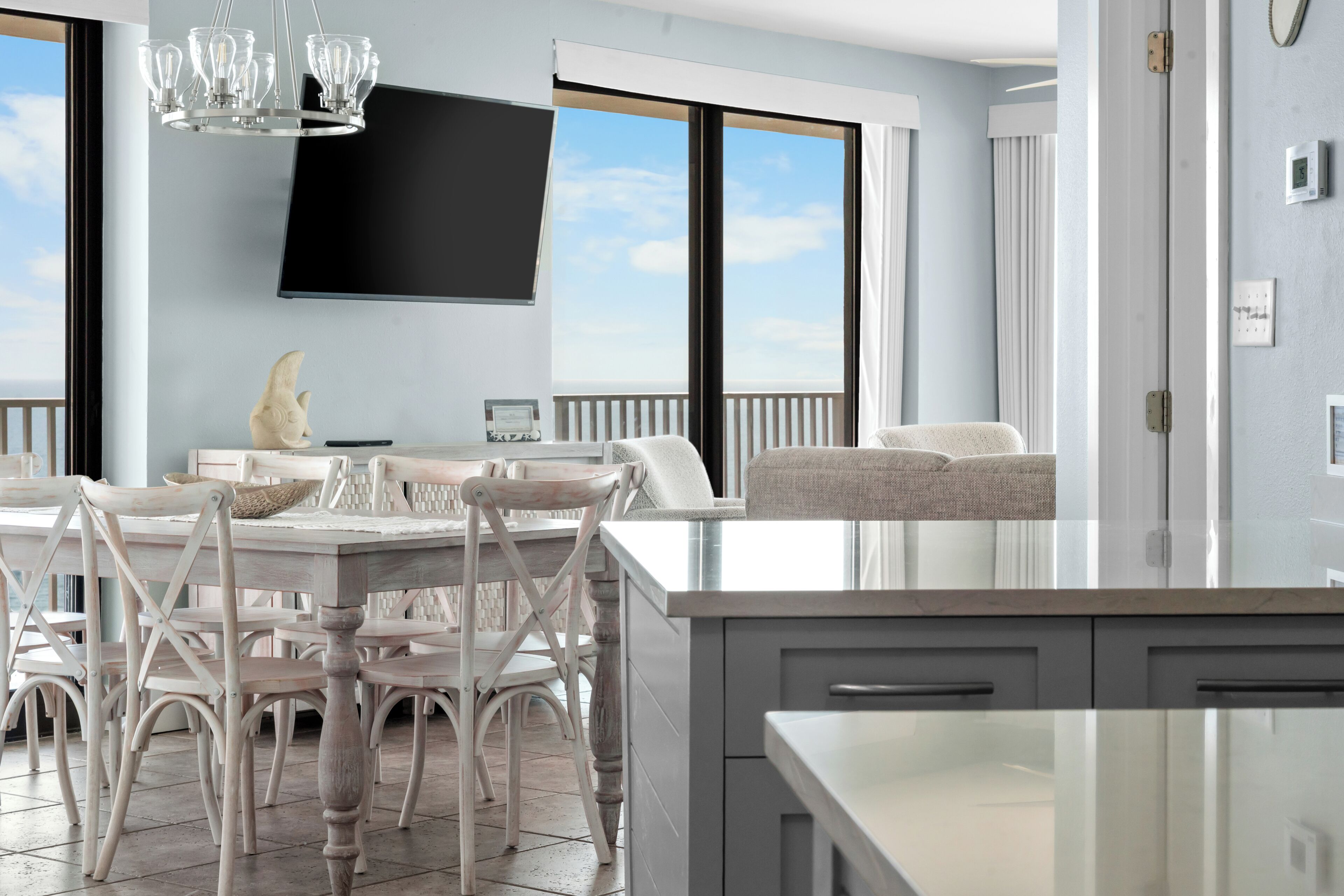 A modern kitchen with a large island, a dining area with white chairs, a flat-screen TV mounted on the wall, and a view of the sky through large windows.