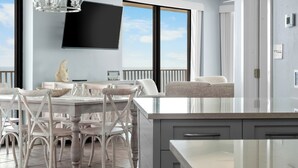 Una cocina moderna con una isla grande, una zona de comedor con sillas blancas, un televisor de pantalla plana montado en la pared y vistas al cielo a través de grandes ventanales.