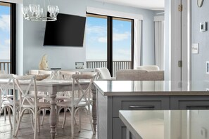 Eine moderne Küche mit einer großen Kochinsel, einem Essbereich mit weißen Stühlen, einem flachbildfernseher an der Wand und Blick zum Himmel durch große Fenster.