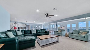 A spacious living area with a green leather sofa, a wooden coffee table, and a grey armchair. A kitchen with white cabinets and a red appliance is visible in the background. - WATERFRONT, PRIVATE POOL, GREAT FOR FAMILES & DOG FRIENDLY! (Gulf Shores)