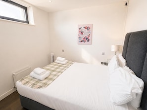 A hotel room with a bed, a window, a radiator, a wall-mounted air conditioner, a bedside lamp, and a floral artwork on the wall.