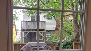 On aperçoit, à travers une porte vitrée, un jardin avec un barbecue abrité, un tapis à rayures bleues et blanches et une terrasse en bois.