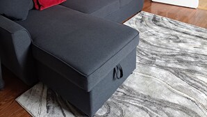 Un salon moderne avec un canapé d’angle gris, un plaid rouge, un tapis à motifs de marbre et un tableau représentant des bateaux colorés accroché au mur.