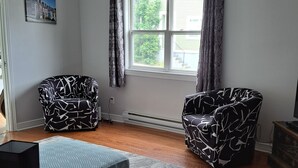 Une pièce comprenant deux fauteuils à motifs, un lit recouvert d’une couverture bleue, une table en bois ornée d’un objet décoratif et une fenêtre avec des rideaux.