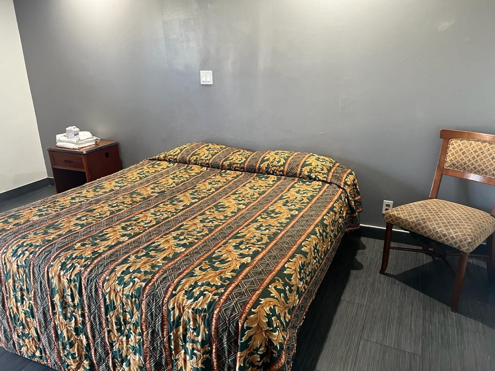 A hotel room with a large bed, a wooden nightstand, a chair, and a gray wall.