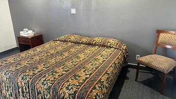 A hotel room with a large bed, a wooden nightstand, a chair, and a gray wall.