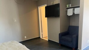 A room with a bed, a dark armchair, a flat-screen TV mounted on the wall, and a door leading to another room. - Pomona Inn (Pomona)