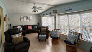 A spacious living room with a ceiling fan, a sofa set, a wicker chair, and a large window with blinds.